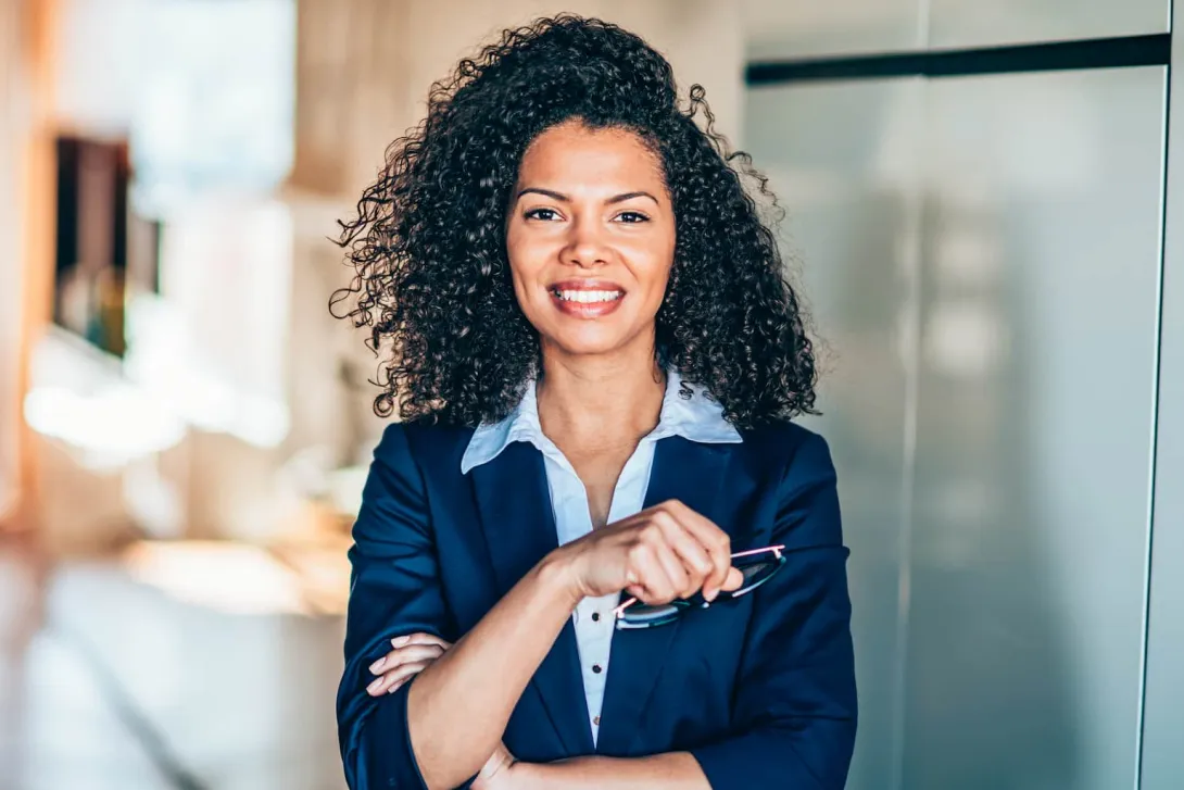 Mulher com cabelo cacheado e sorriso confiante, usando um terno azul, segurando um óculos com as duas mãos, em um ambiente de escritório moderno e iluminado.